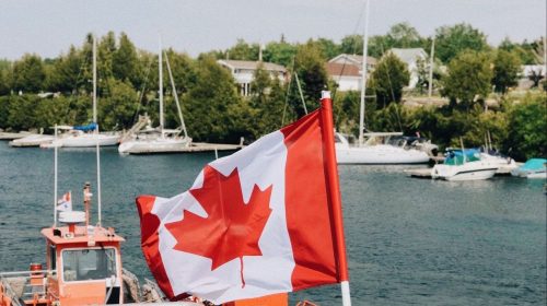 A vibrant Canadian flag waving at a harbor with boats nearby during a summer day.